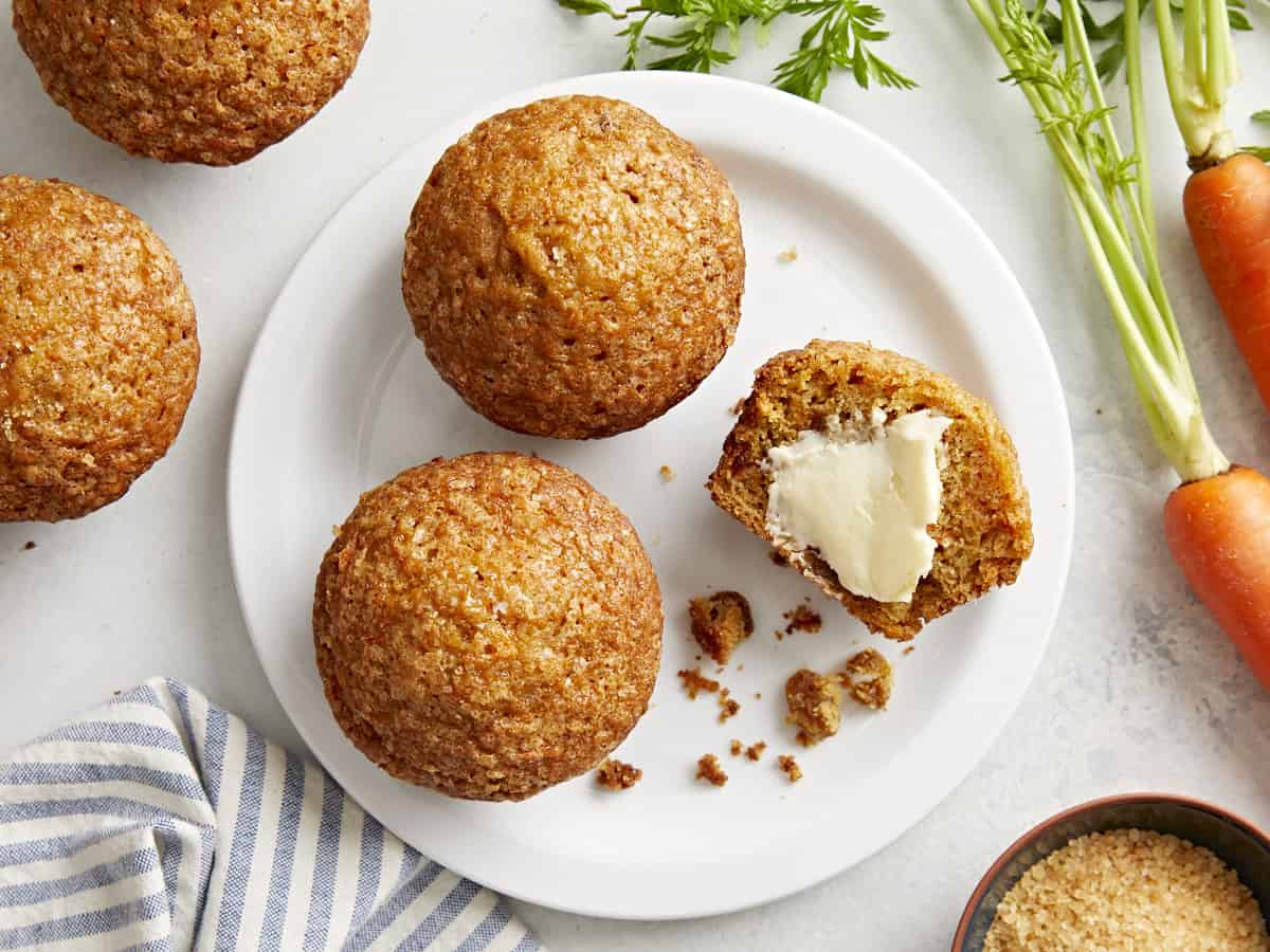 Top view of three carrot cake muffins on a white plate and two carrot cake muffins lying next to the plate.