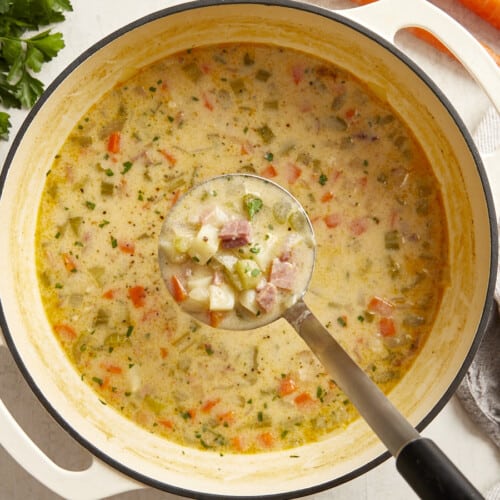 a ladle full of ham and potato soup hanging over a pot of soup.