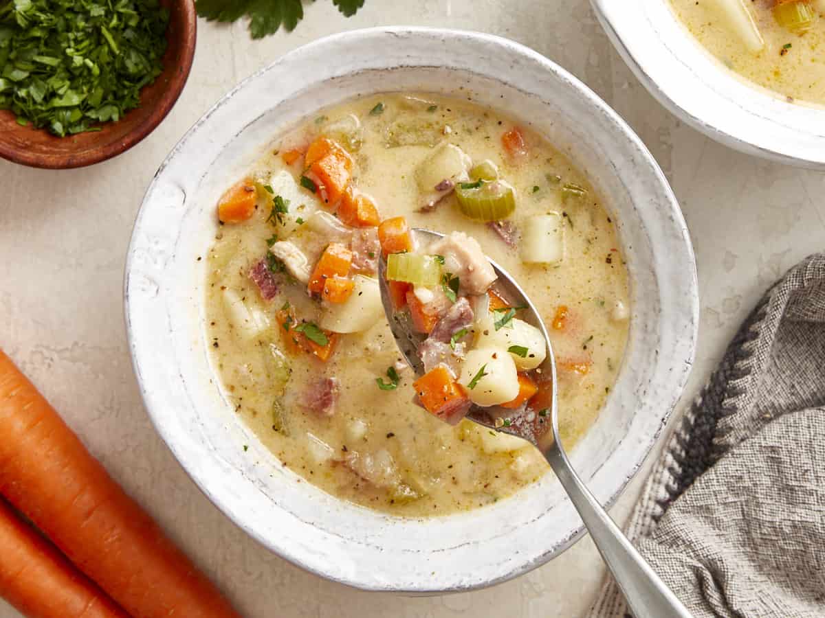 Top view of a spoonful of ham and potato soup hanging over a portion of soup in a white bowl.