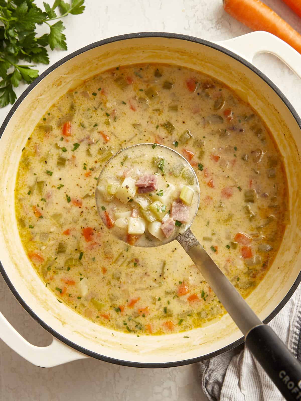 a ladle full of ham and potato soup hanging over a pot of soup.