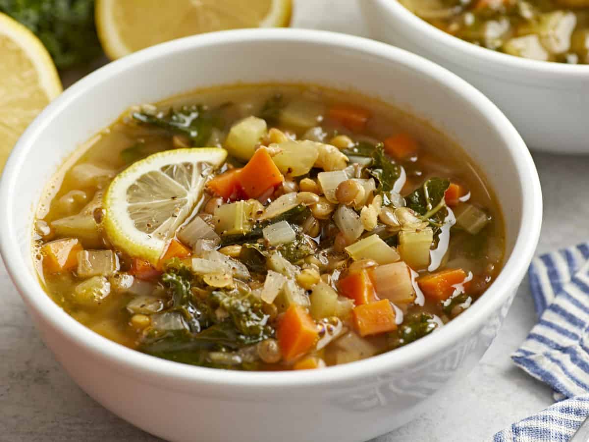 Three-quarter view of a Mediterranean lentil soup in a white bowl with a slice of lemon.