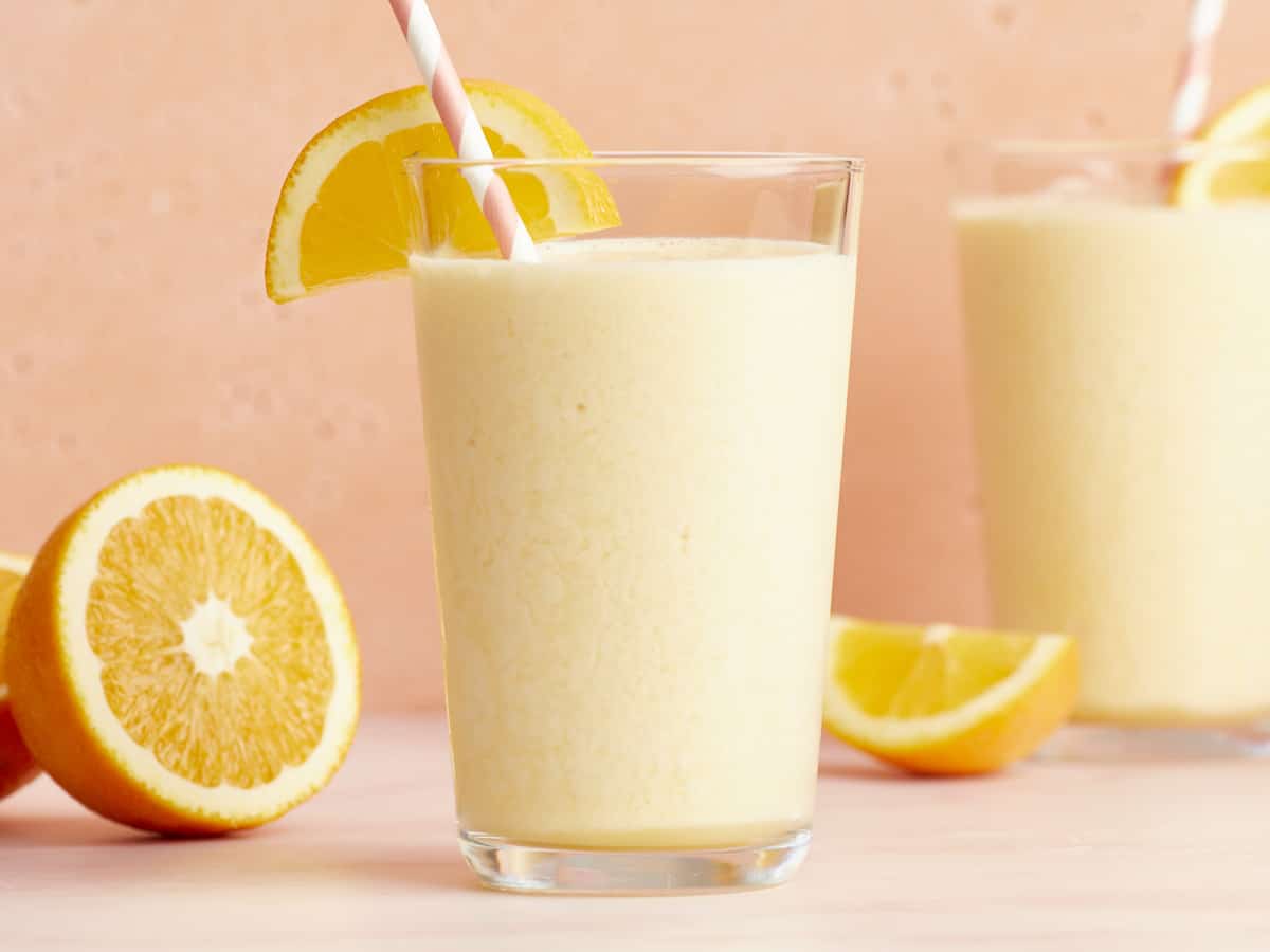 Side view of two glasses full of Orange Julius with striped straws and orange slices.