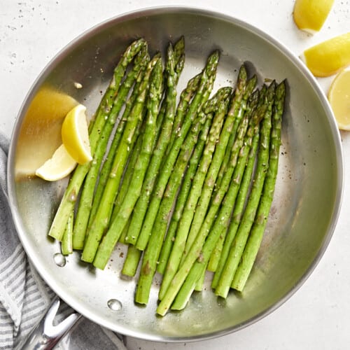 Top view of sautéed asparagus in a skillet with lemon wedges.