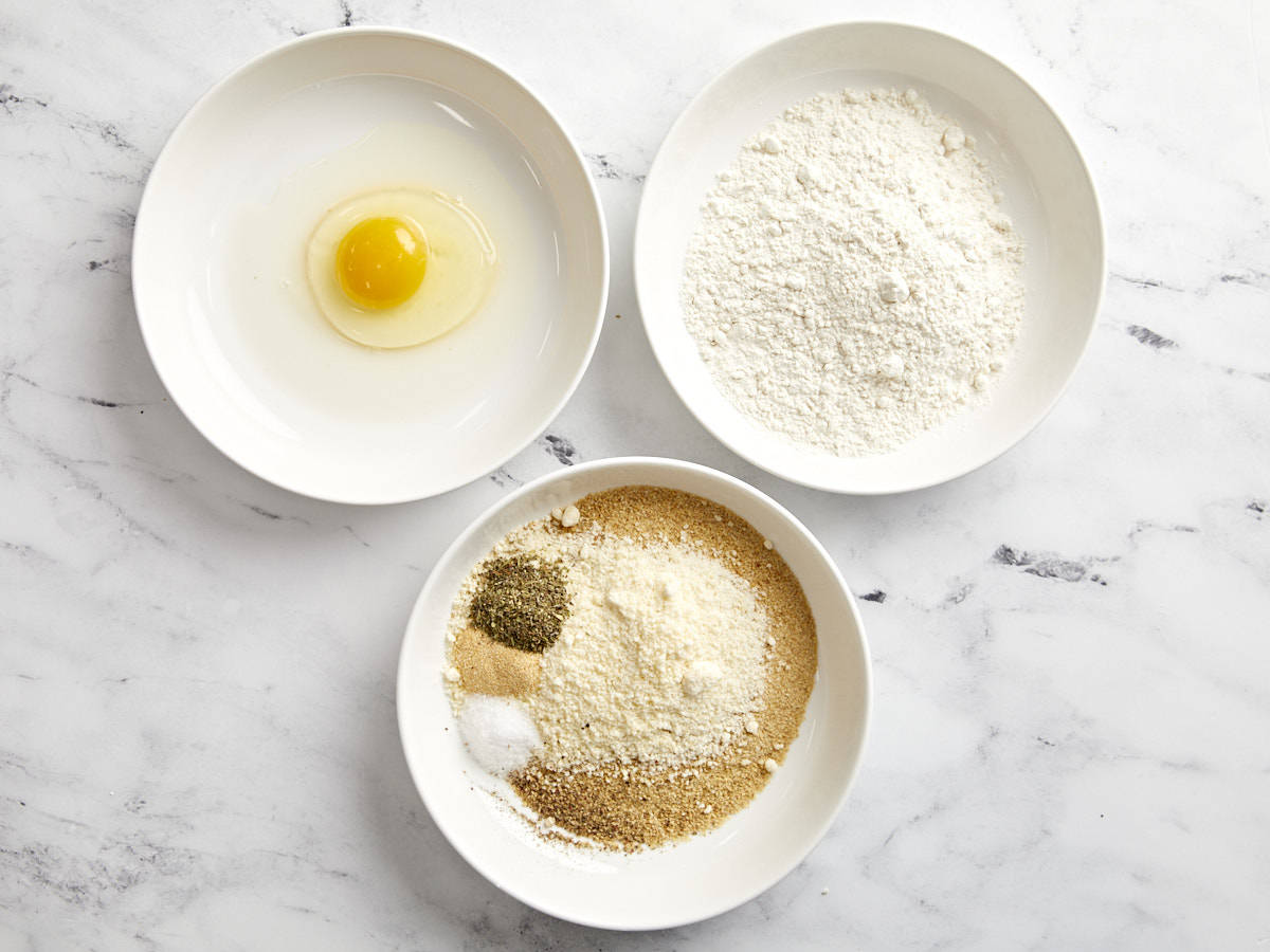 an egg, flour, and seasoned breadcrumbs in separate white bowls.