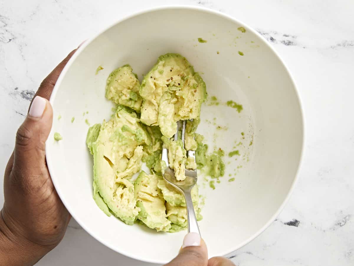 A hand holding a fork and mashing avocado in a white bowl.