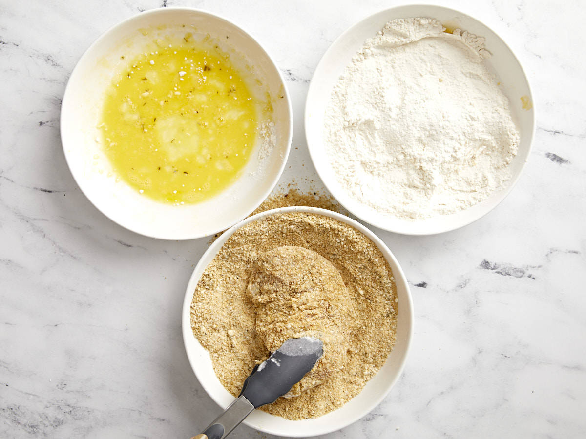 dredging a chicken breast through egg, flour, and breadcrumbs in separate bowls.