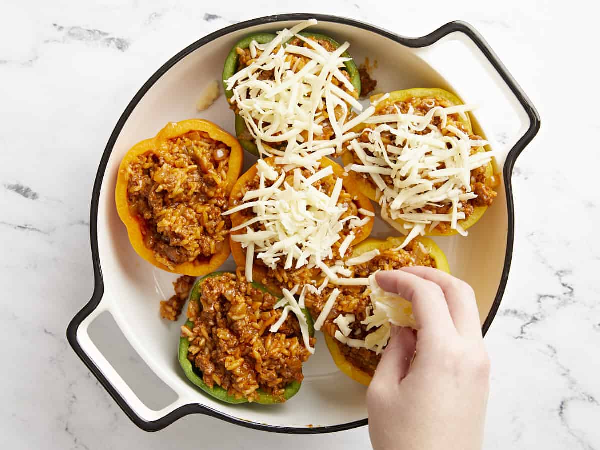 Filled bell peppers in a baking dish being topped with shredded cheese.