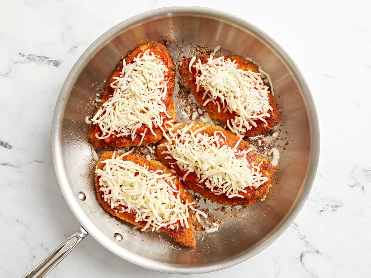 4 fried chicken breasts topped with marinara sauce and shredded cheese in a stainless skillet.