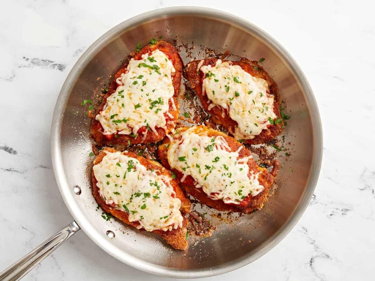 parsley topped fried chicken breasts with marinara sauce and melted cheese in a stainless skillet.