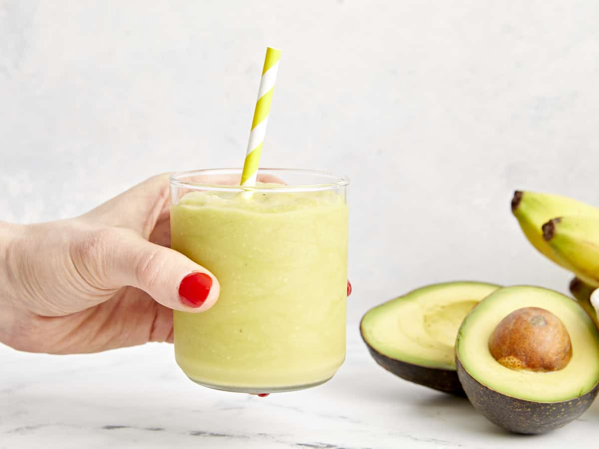 a hand holding an avocado smoothie in a clear glass with a straw.