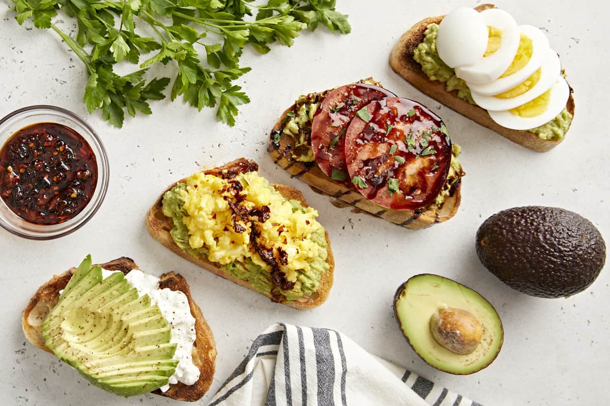 Top view of avocado toast prepared 4 different ways, with parsley, avocado and a napkin on the side.