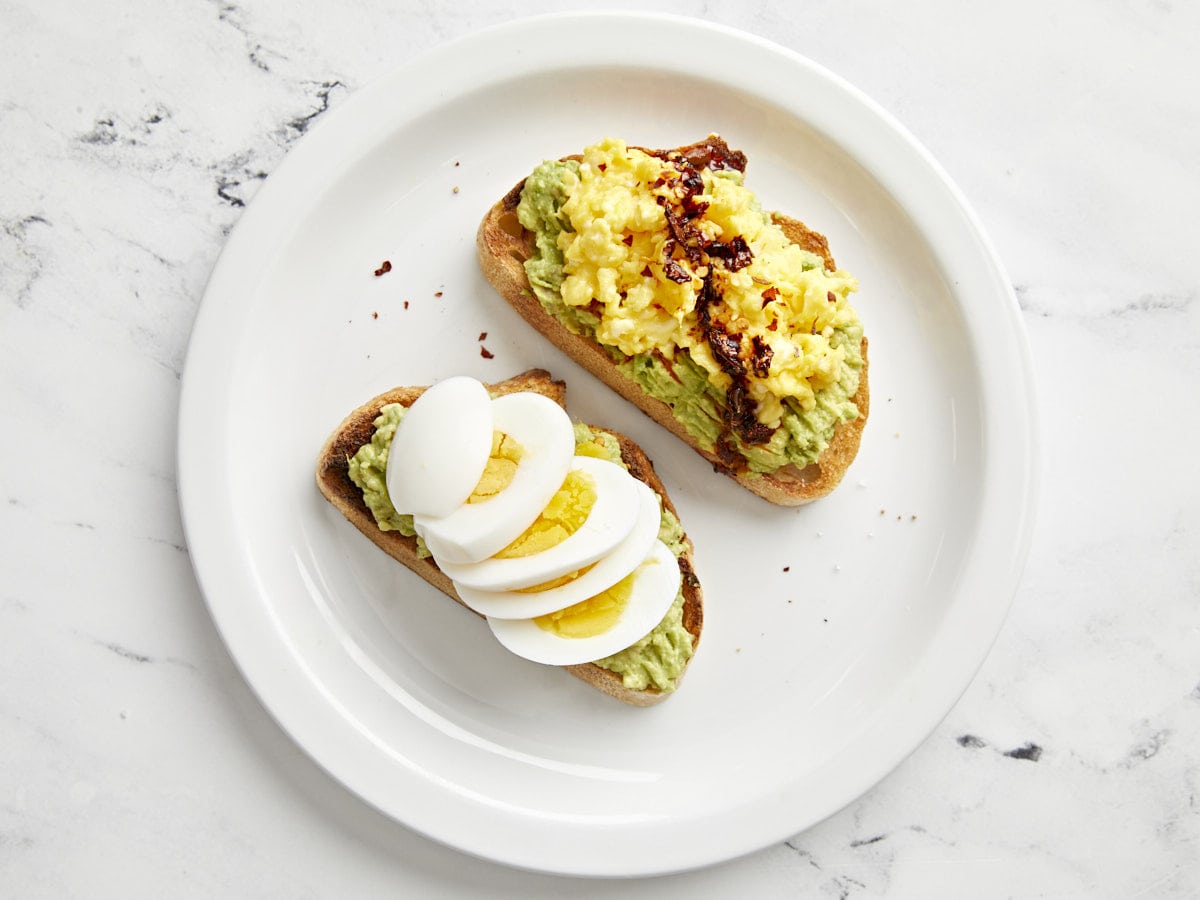 Two slices of avocado toast with a hard boiled egg and soft scrambled eggs on top.