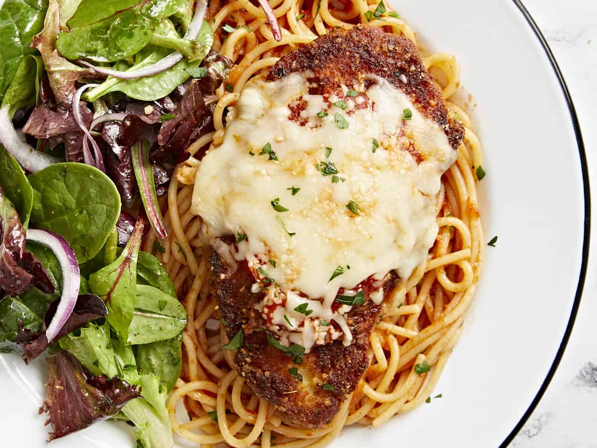 close up overhead view of a serving of chicken parmesan over spaghetti with a side salad on a white plate.