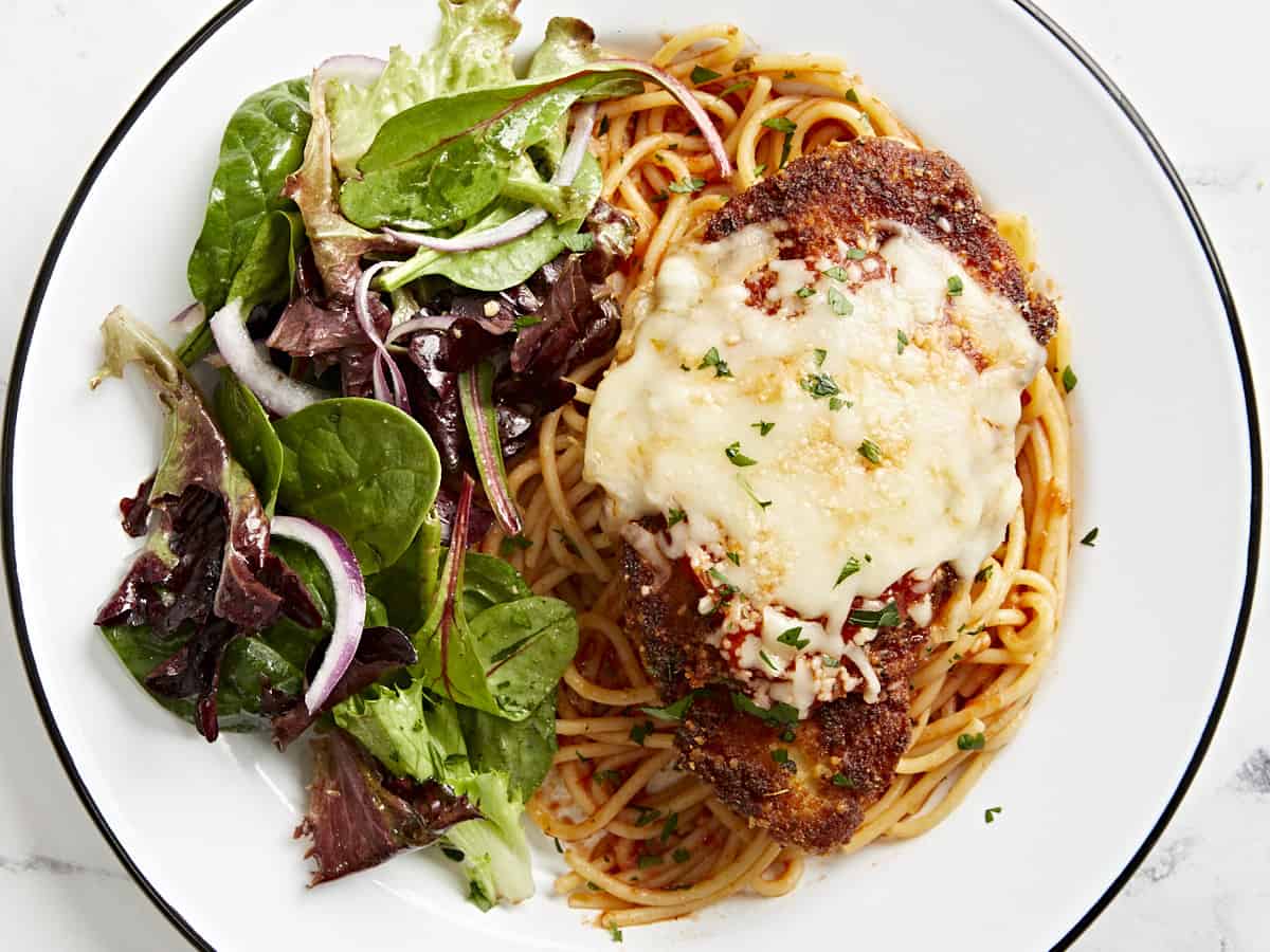 overhead view of a serving of chicken parmesan over spaghetti with a side salad on a white plate.