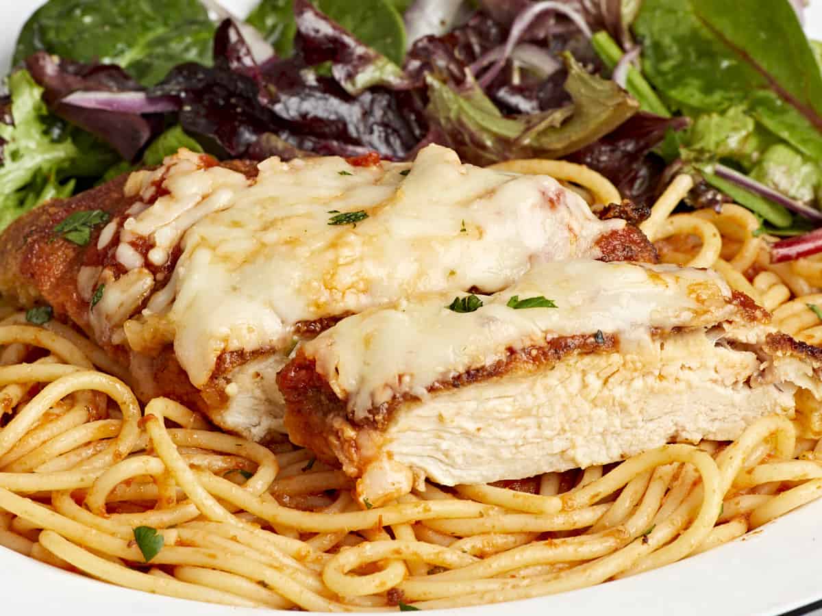 side view of a sliced serving of chicken parmesan on a bed of spaghetti with a side salad.