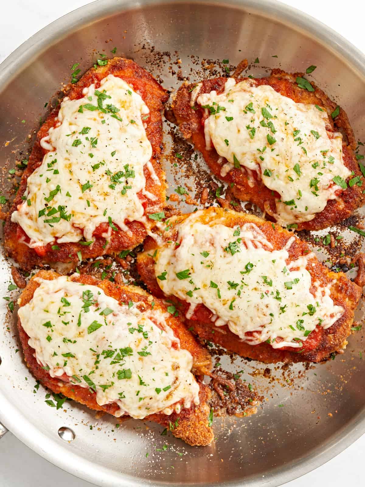 overhead view of 4 fried chicken breasts topped with marinara sauce, melted cheese, and parsley in a stainless skillet.