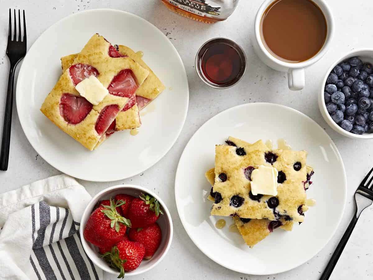 Overhead view of sliced sheet pan pancakes on two serving dishes with fruit and syrup on the side.