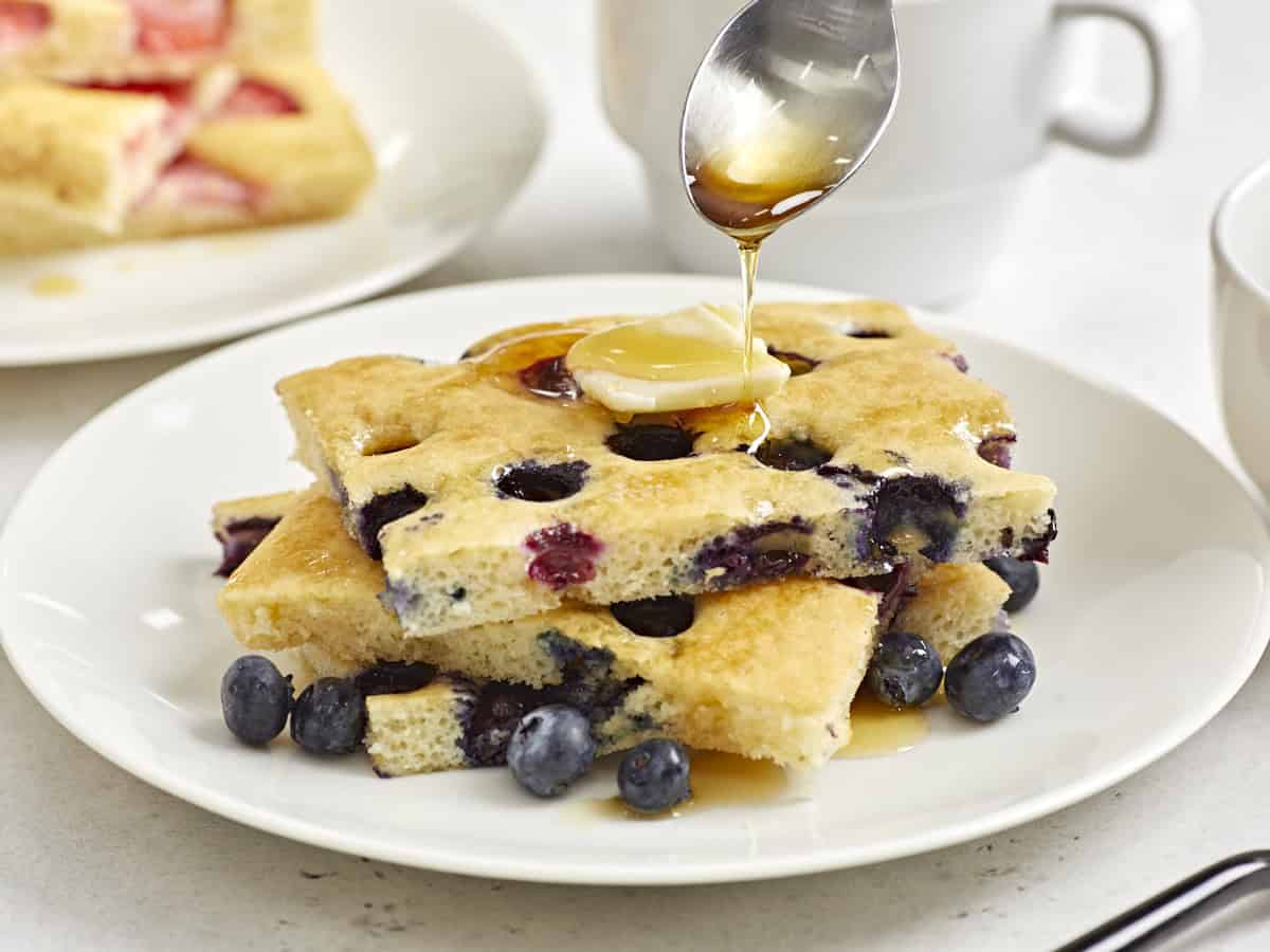 Close up side view of sheet pan pancakes on a serving plate with maple syrup being drizzled over the top.