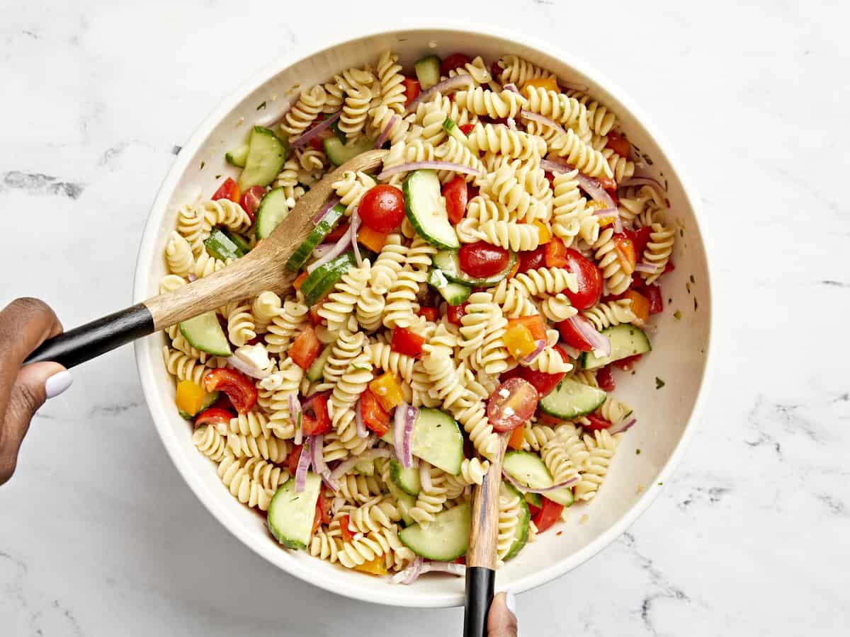 Pasta salad being tossed and mixed together with serving utensils.
