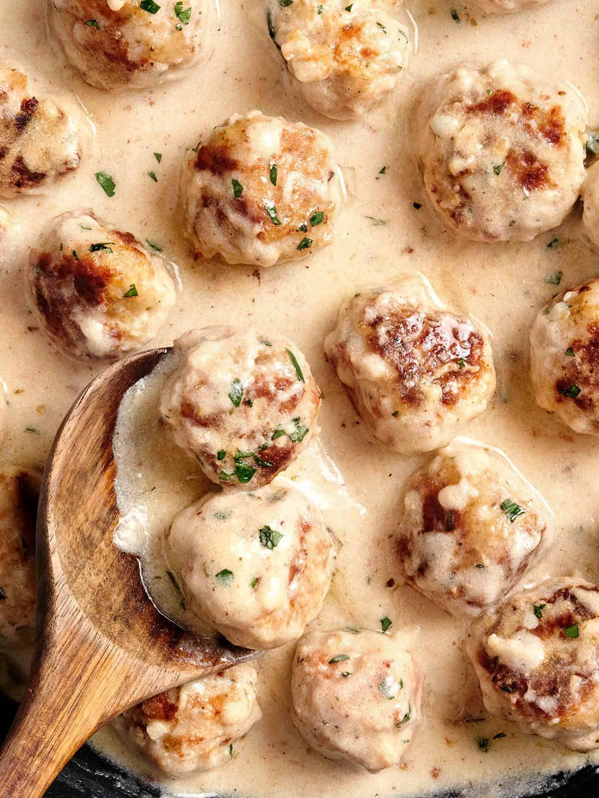 Close up of chicken meatballs in cream sauce in a skillet with a wooden spoon.