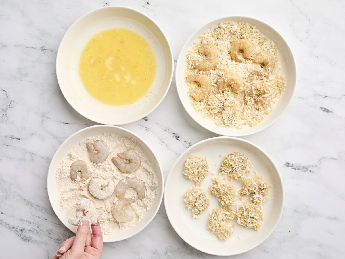 Shrimp being dredged in egg wash, then coconut and panko breadcrumbs, and then flour.