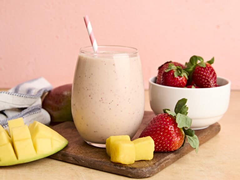A glass of homemade strawberry mango smoothie with a straw, on a wooden cutting board with strawberries and mango chunks.