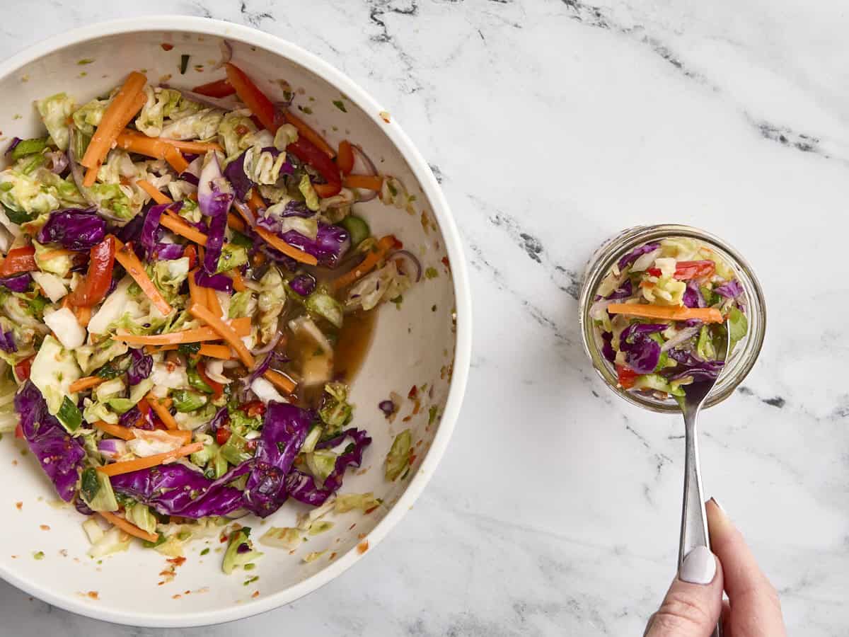 Prepped and massaged kimchi vegetables in a mixing bowl, with a spoon portioning some into a glass mason jar.