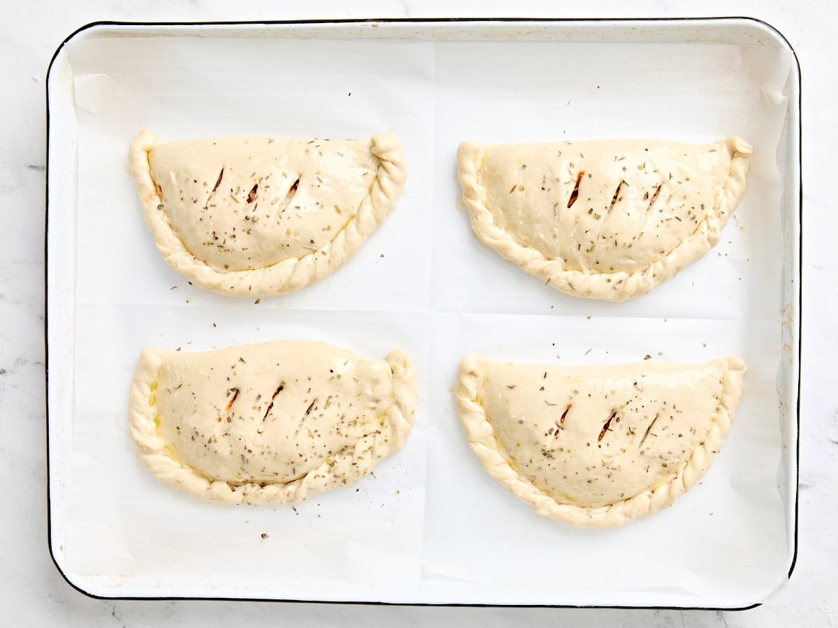Four calzones on a parchment lined baking sheet with 3 slits cut in the top before baking.