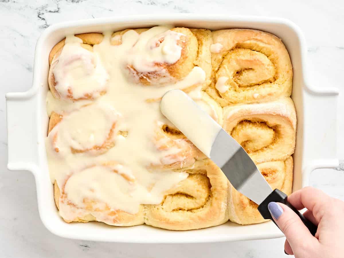 An offset spatula spreading cream cheese frosting over homemade orange rolls.