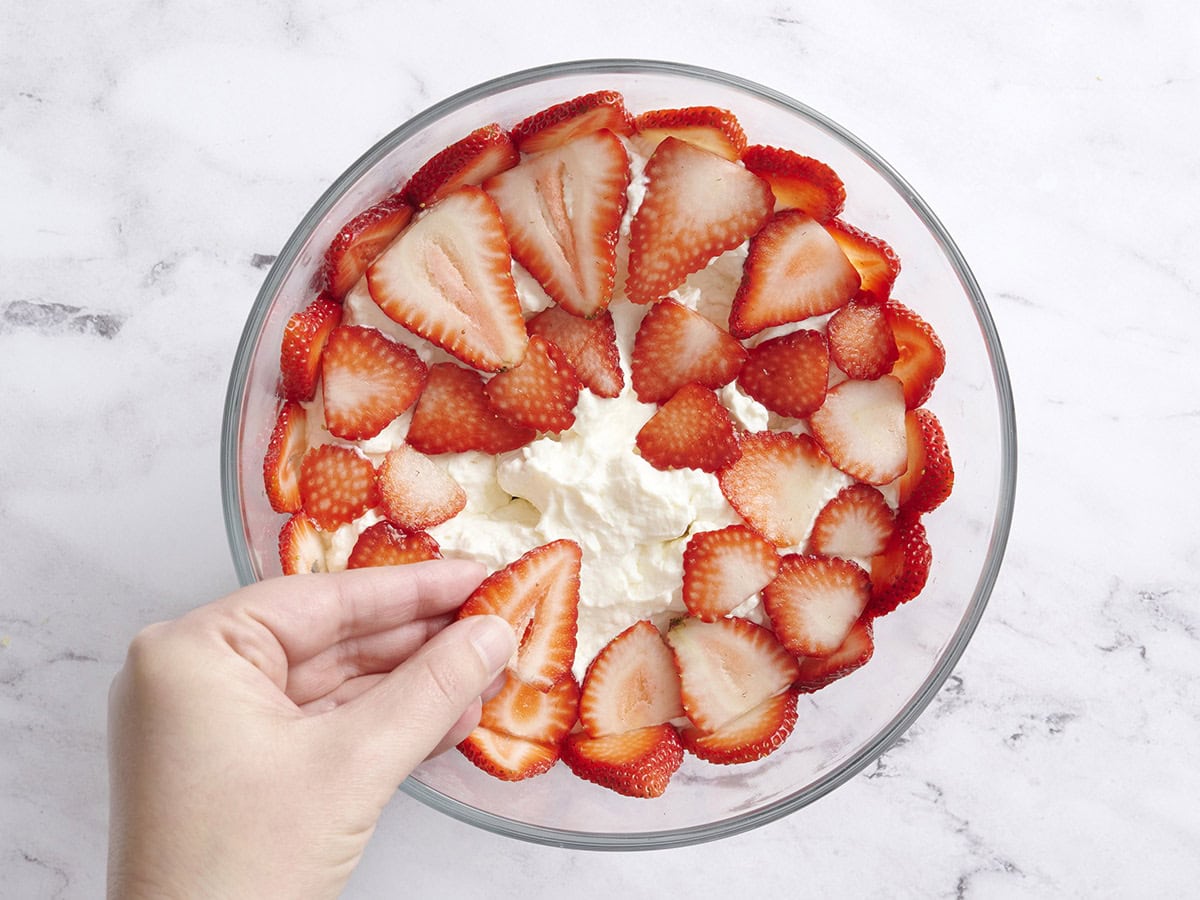 A hand placing slices of strawberries on a blue white and red trifle.
