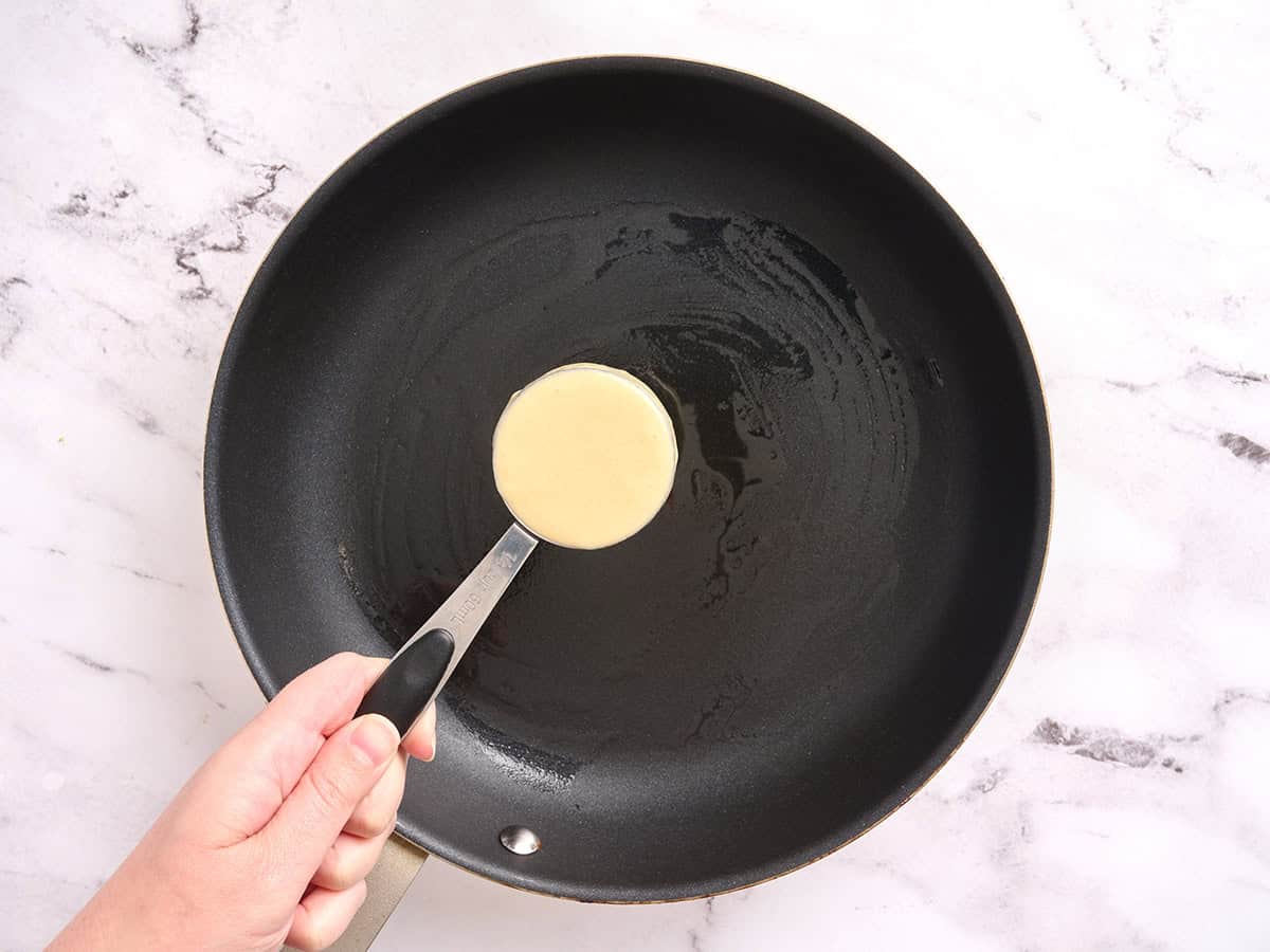 Pancake batter in a cup measure being held over a skillet.
