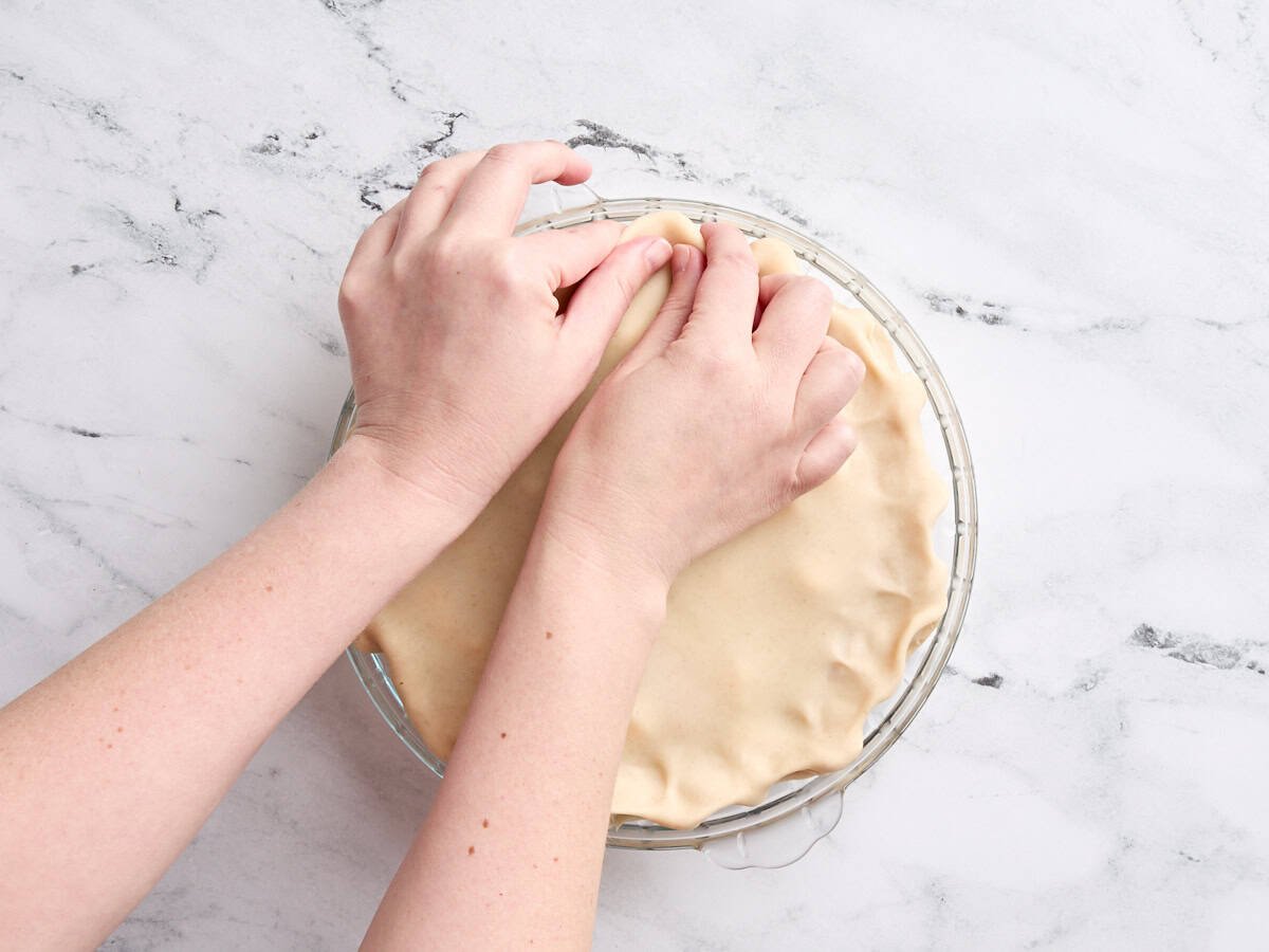 Hands crimping the edges of a pie crust in a pie dish.