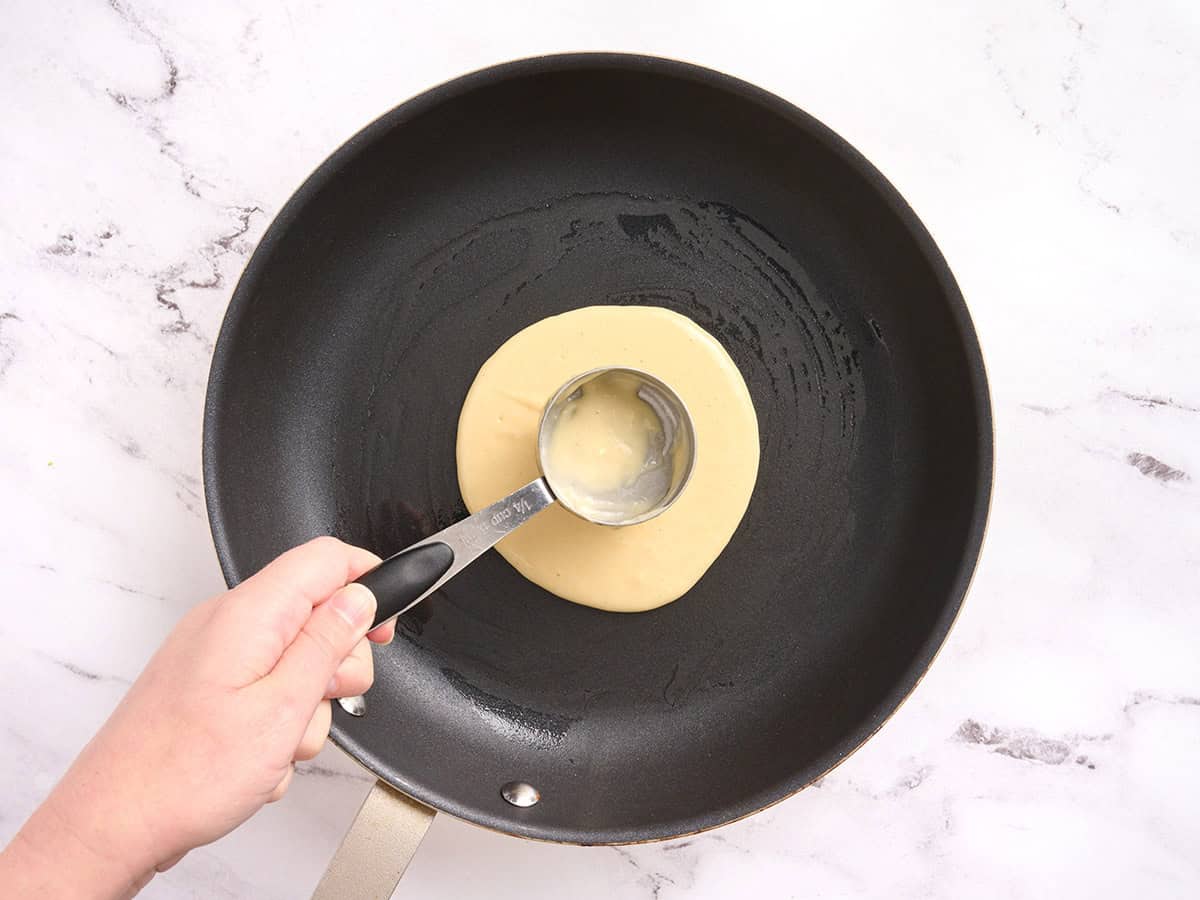A measuring cup flattening pancake batter in a skillet.