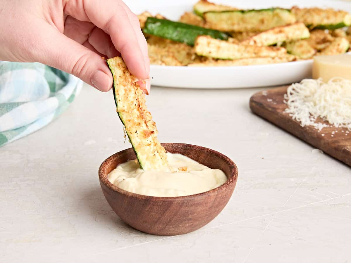 An air fried zucchini spear being dipped into a bowl of mayonnaise. 