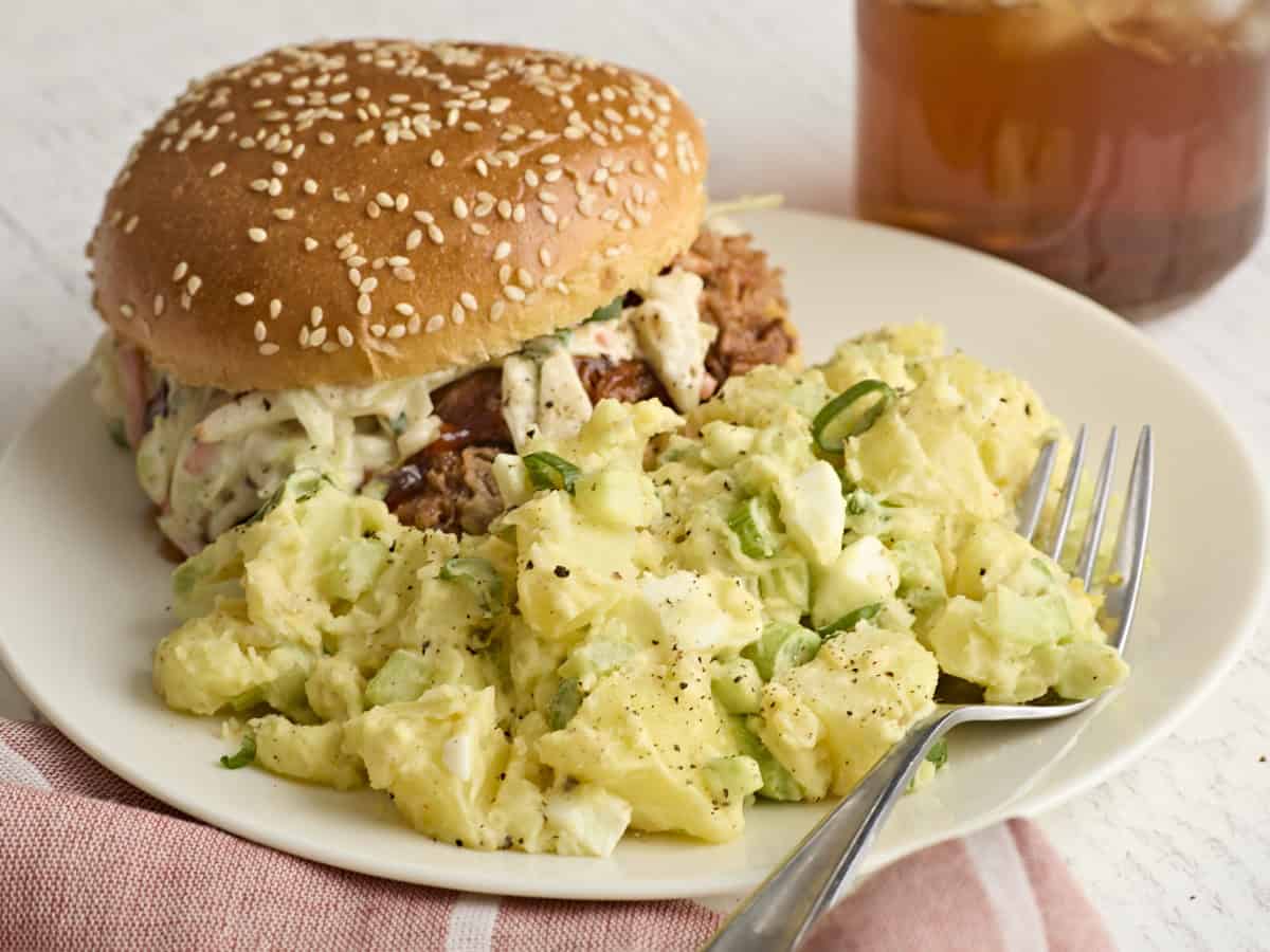 Side view of creamy potato salad on a plate with a pulled pork sandwich.