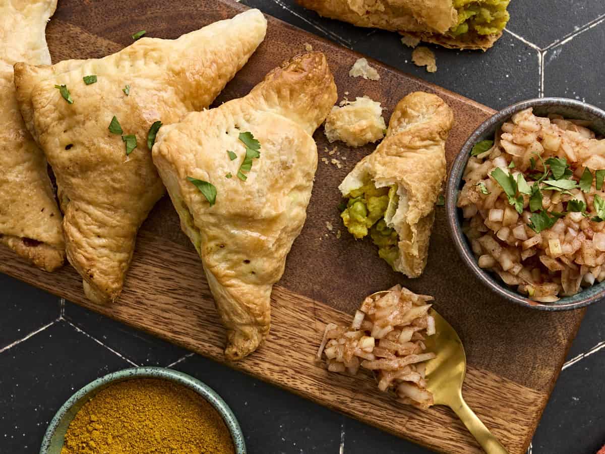 Overhead view of homemade puff pastry samosas on a wooden board with a bowl of onion chutney.