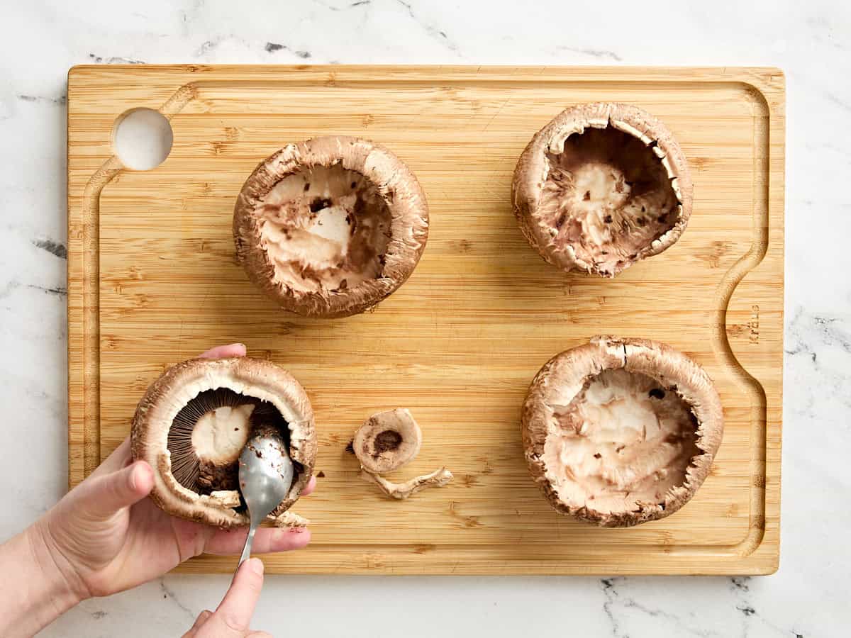 A spoon scooping out the gills from four portabella mushrooms on a wooden cutting board.