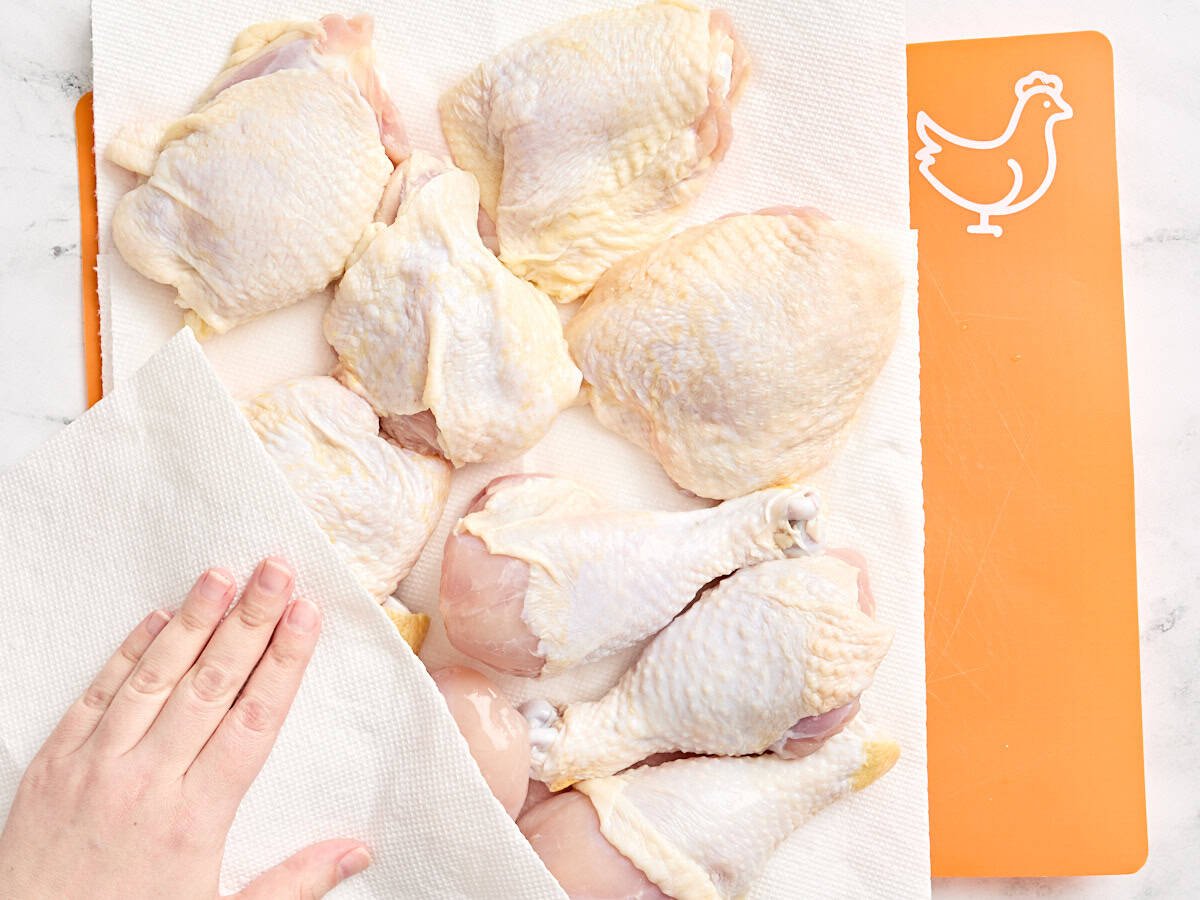 A hand using a paper towel to pat chicken drumsticks and thighs dry.