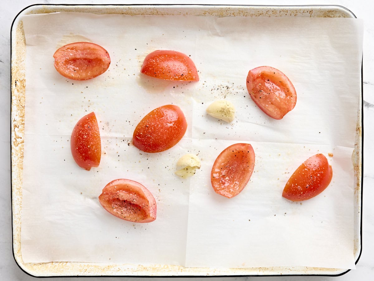 A cut up deseeded tomato and crushed garlic cloves on a baking sheet.