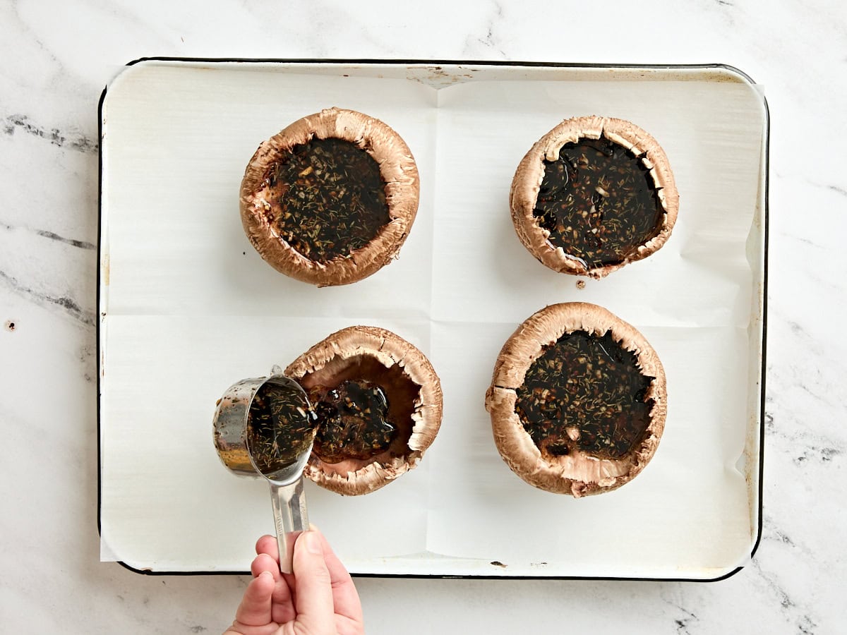 A spoon drizzling a balsamic marinade over four portabella mushrooms on a parchment lined baking sheet.