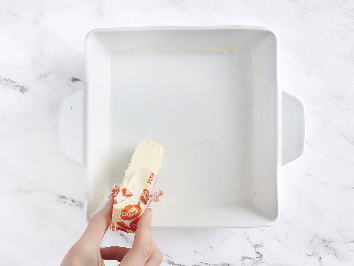 Hands using a stick of butter to grease a baking dish.