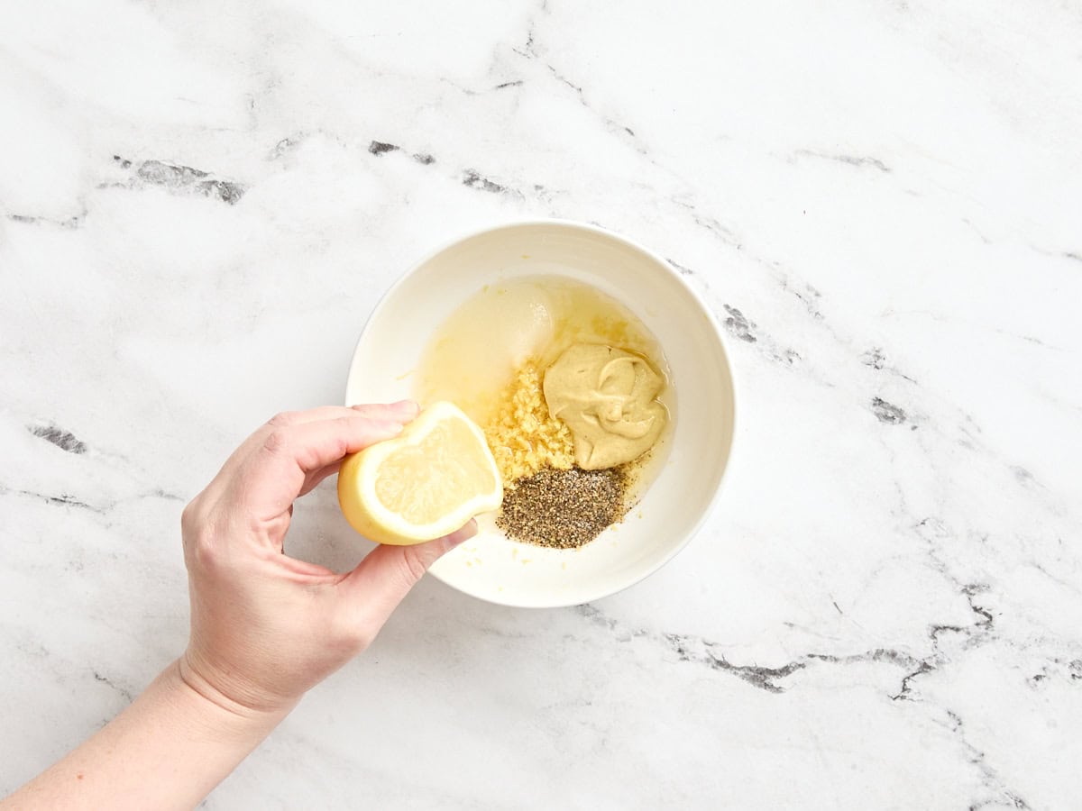A hand squeezing half a lemon into a bowl of apple cider vinegar, mustard, sugar and seasonings.