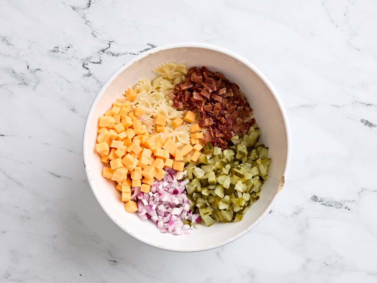 Cooked bowtie pasta, cubed cheddar, cooked and crumbled bacon, diced dill pickles. and diced red onion in a bowl.