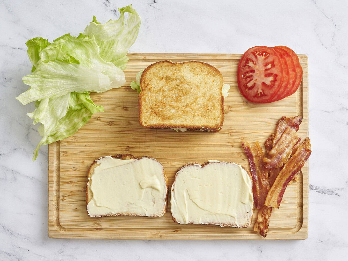 Overhead view of two slices of toasted bread topped with mayo, cooked bacon, lettuce, sliced tomato and a completed BLT sandwich