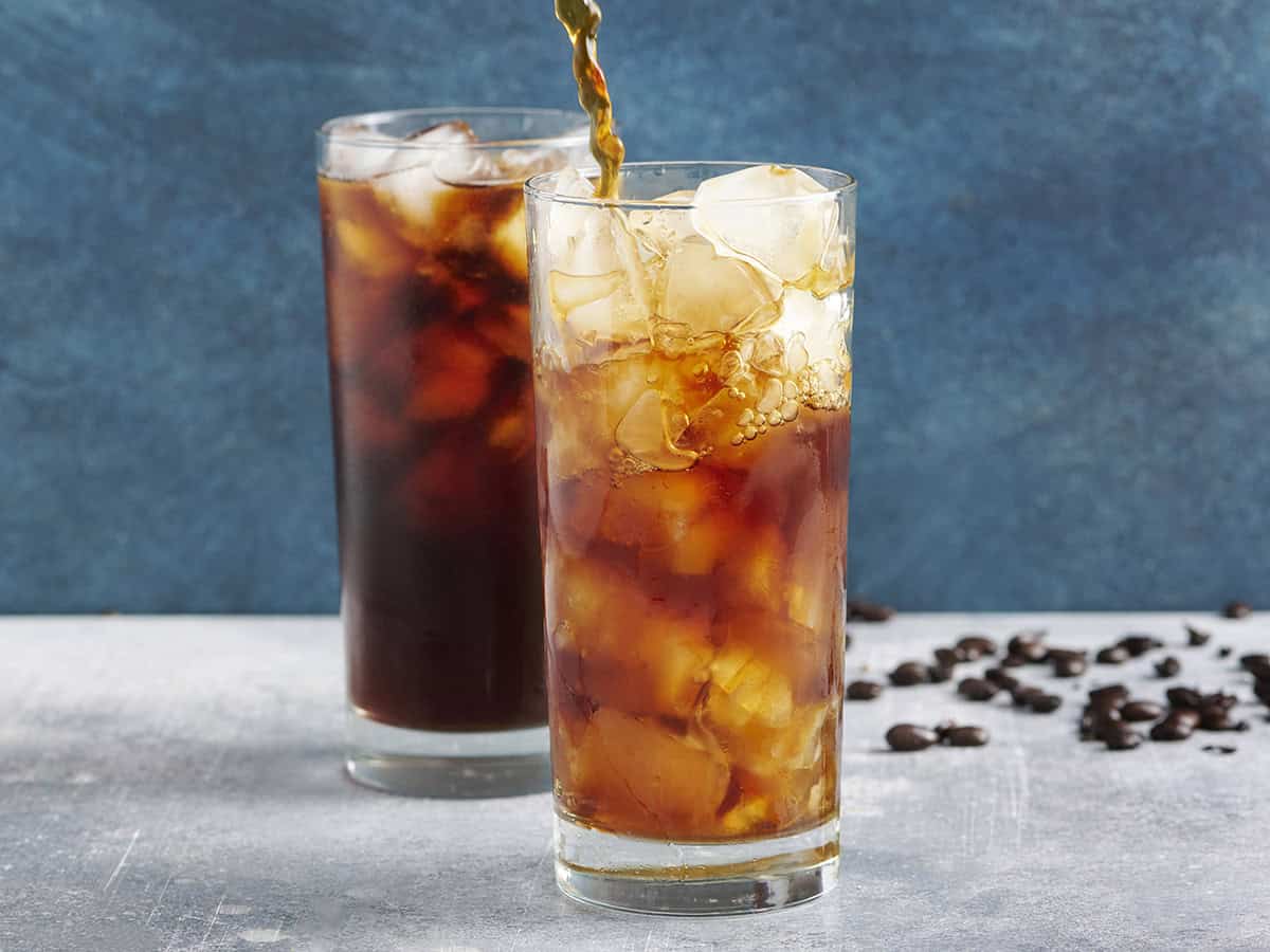 Side view of two glassed of cold brew coffee with ice, with the coffee being poured into one of the glasses.
