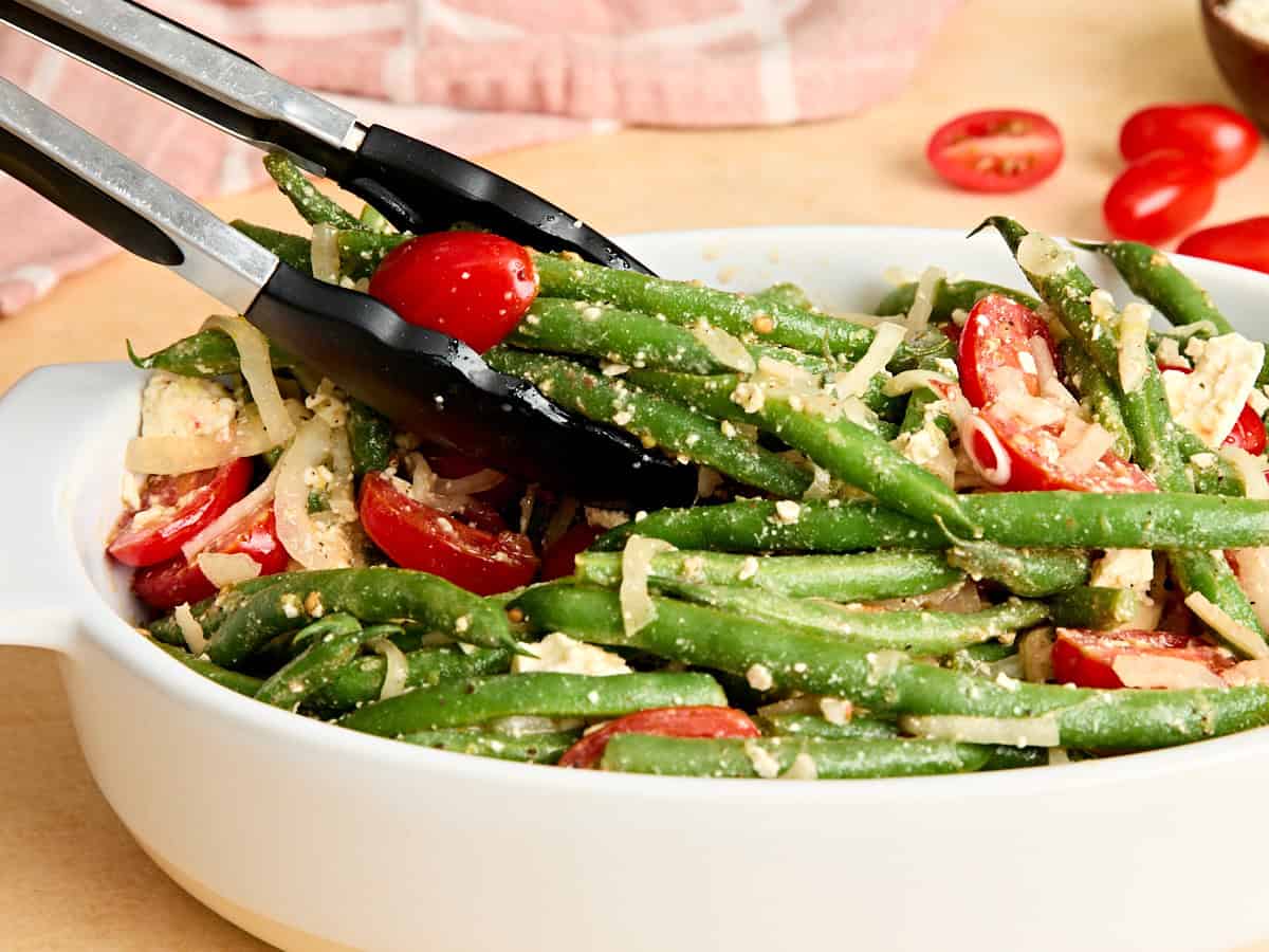 Side view of kitchen tongs taking some green bean salad from a dish.