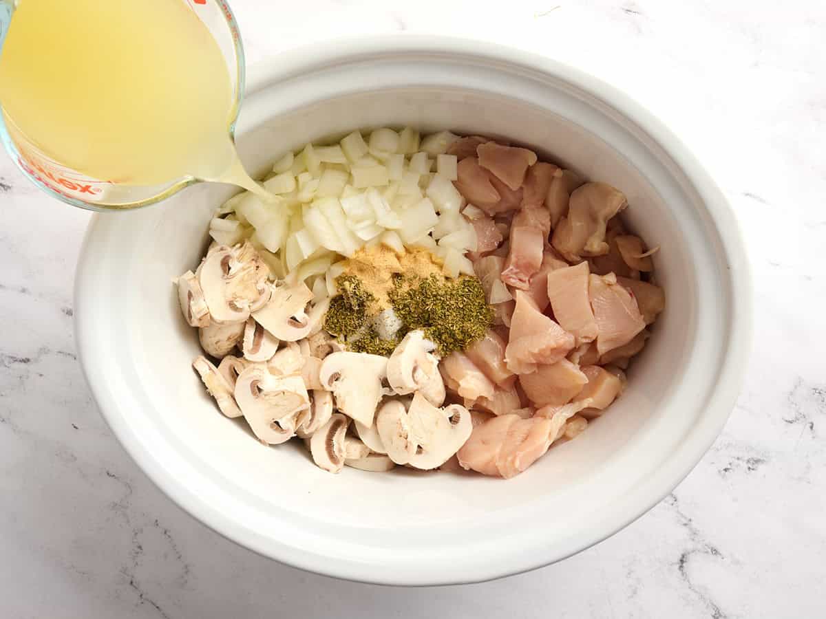 Raw diced chicken breast, sliced mushrooms, diced onion, and seasonings in a crockpot with broth being poured on top.