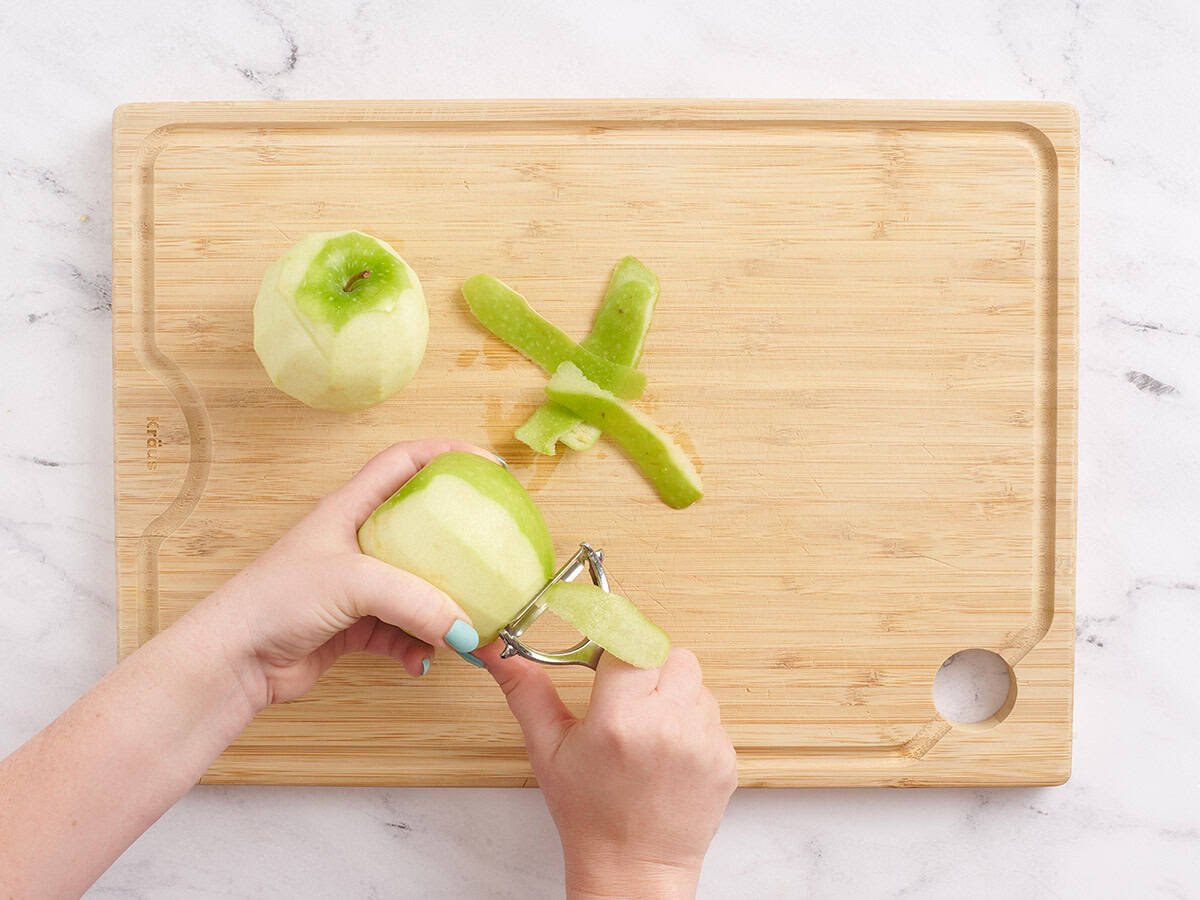 A hand using a vegetable peeler to peel a green apple.