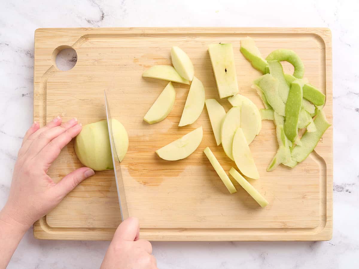 Hands slicing a peeled apple, with the apple peel discarded to the side on a wooden cutting board.
