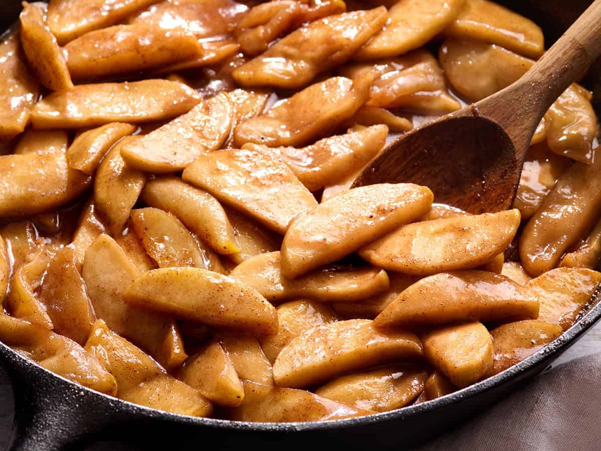 Side view of a skillet of fried apples with a wooden spoon stirring them.